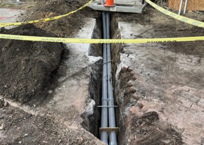 A trench containing metal pipes is sectioned off with caution tape and a traffic cone. In the background are buildings, vehicles, and construction materials.